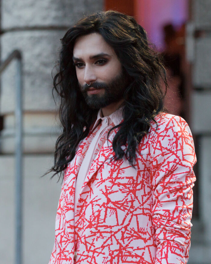 Conchita Wurst with long black hair and beard, wearing a patterned red and white jacket outdoors near a building.