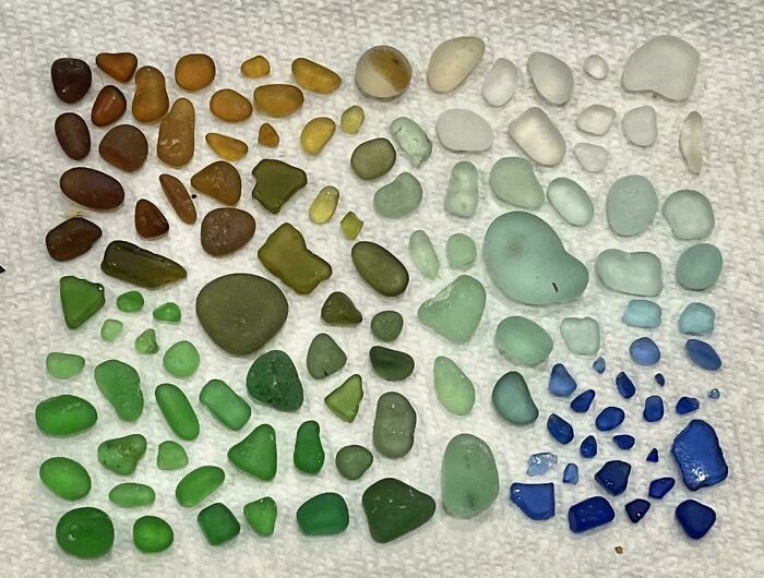 Colorful sea glass pieces neatly arranged by shade and size on a white textured surface, showcasing knolled order.