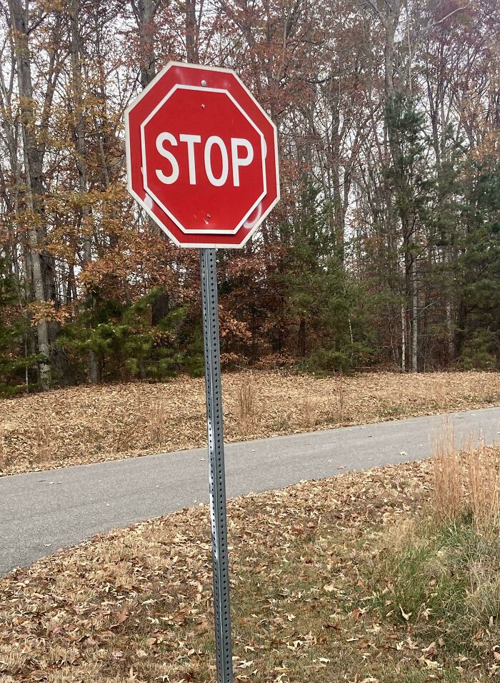 Stop sign mounted crookedly on a bent pole beside a quiet road, illustrating people not even trying in maintenance.