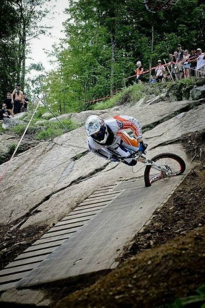 Mountain biker frozen in action on a steep rocky trail surrounded by spectators and dense forest foliage.