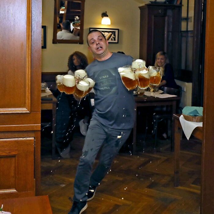 Man carrying multiple foamy beer glasses in a lively bar, a frozen frame capturing the moment before a spill.