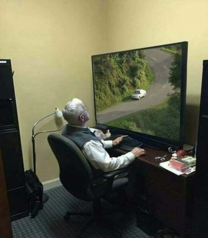 Elderly man sitting at desk watching a large screen displaying a frozen frame of a car on a winding road.