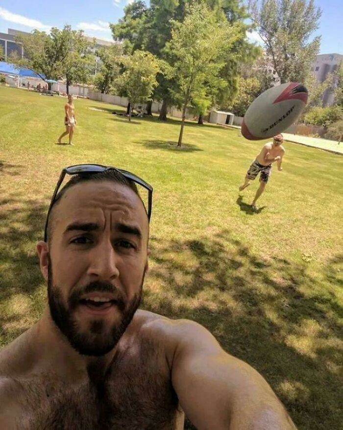 Man taking a selfie with a rugby ball flying toward him outdoors, capturing a frozen frame moment without showing aftermath.