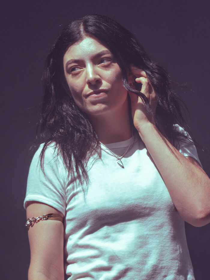Lorde in a white t-shirt adjusting her hair while looking to the side during a performance or event.