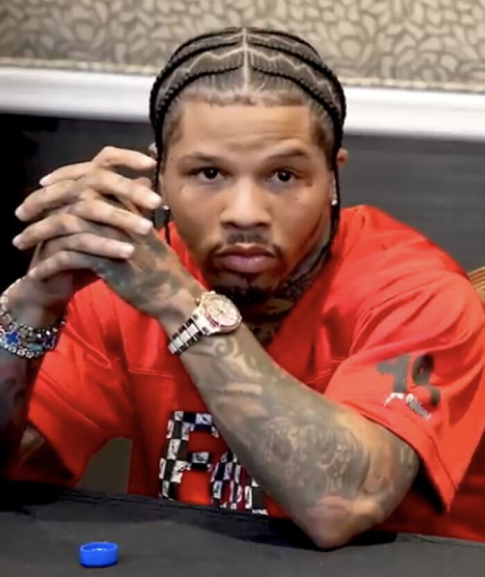 Gervonta Davis with braided hair and tattoos, wearing a red shirt and watch, sitting at a table in a casual setting