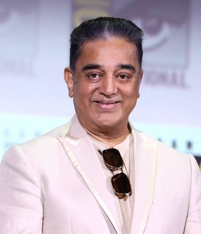 Kamal Haasan smiling, wearing a cream blazer with sunglasses hanging on the collar at a public event.
