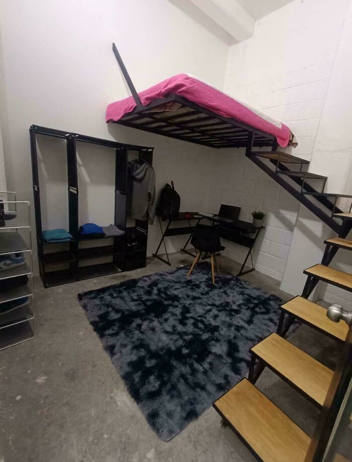 Loft bed accessed by steep stairs in a compact Airbnb room with minimalist furniture and a dark gray shaggy rug.