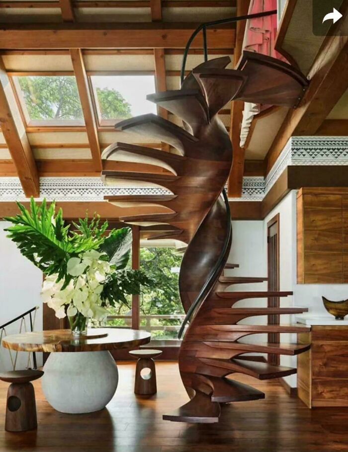 Spiral wooden staircase with a modern design inside a sunlit room featuring wooden furniture and large plant.