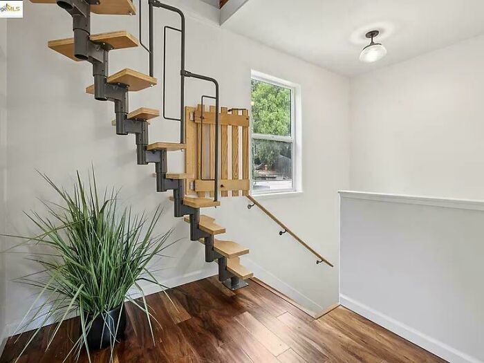 Spiral wooden stairs with black metal handrails in a bright Airbnb rental interior with hardwood floors and a potted plant.