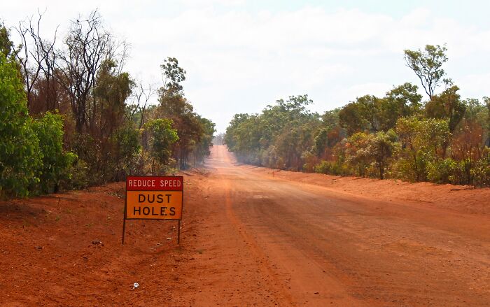 Red dirt remote road with trees on both sides and a warning sign about dust holes in a remote location.