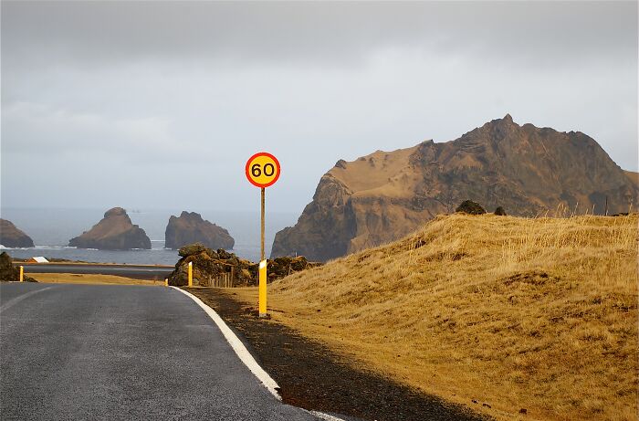 Remote locations with winding road, speed limit sign, barren hills, and rocky coastal cliffs under cloudy sky.