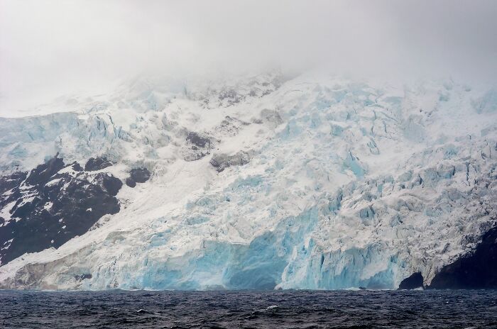 Glacier-covered remote location with icy cliffs and dark ocean waters under a misty, overcast sky.