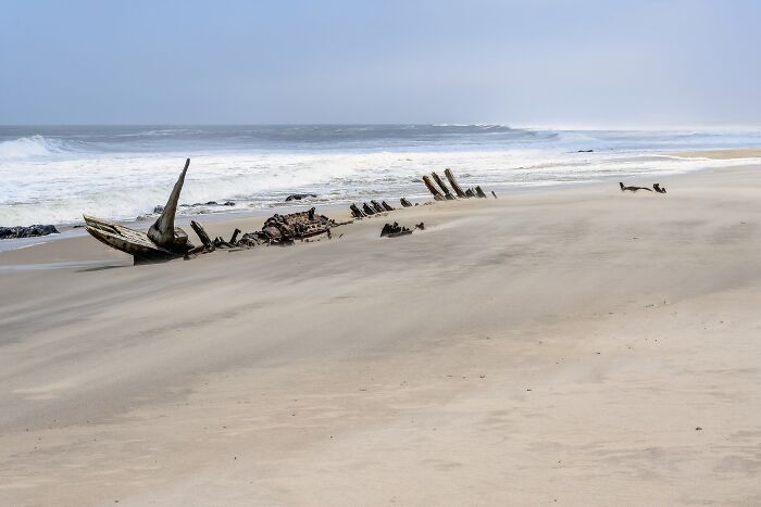 Rusty shipwreck on a vast sandy beach at a remote location with ocean waves under a clear sky.