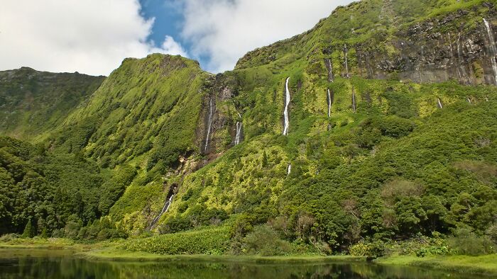 Lush green cliffs with multiple waterfalls in a remote location surrounded by dense vegetation and a calm water body.