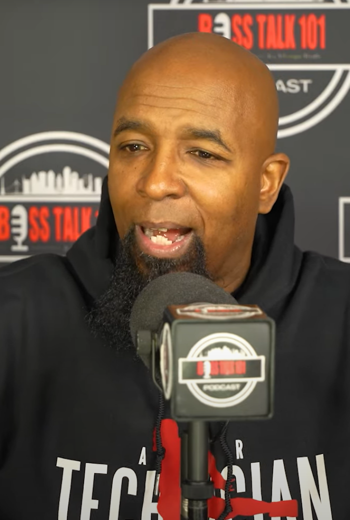 Tech N9ne speaking into a podcast microphone, wearing a black hoodie with Tech N9ne logo on it.