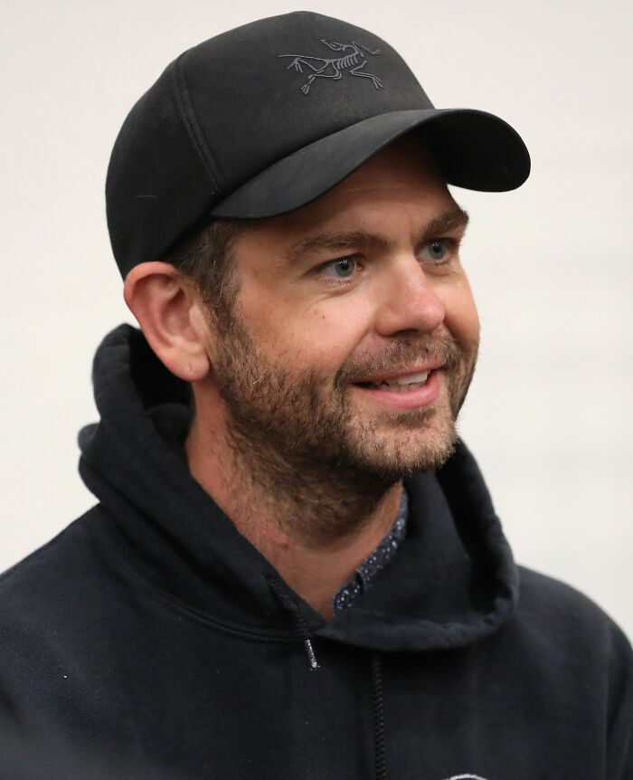 Jack Osbourne wearing a black cap and hoodie, smiling and looking to the side in a casual indoor setting.