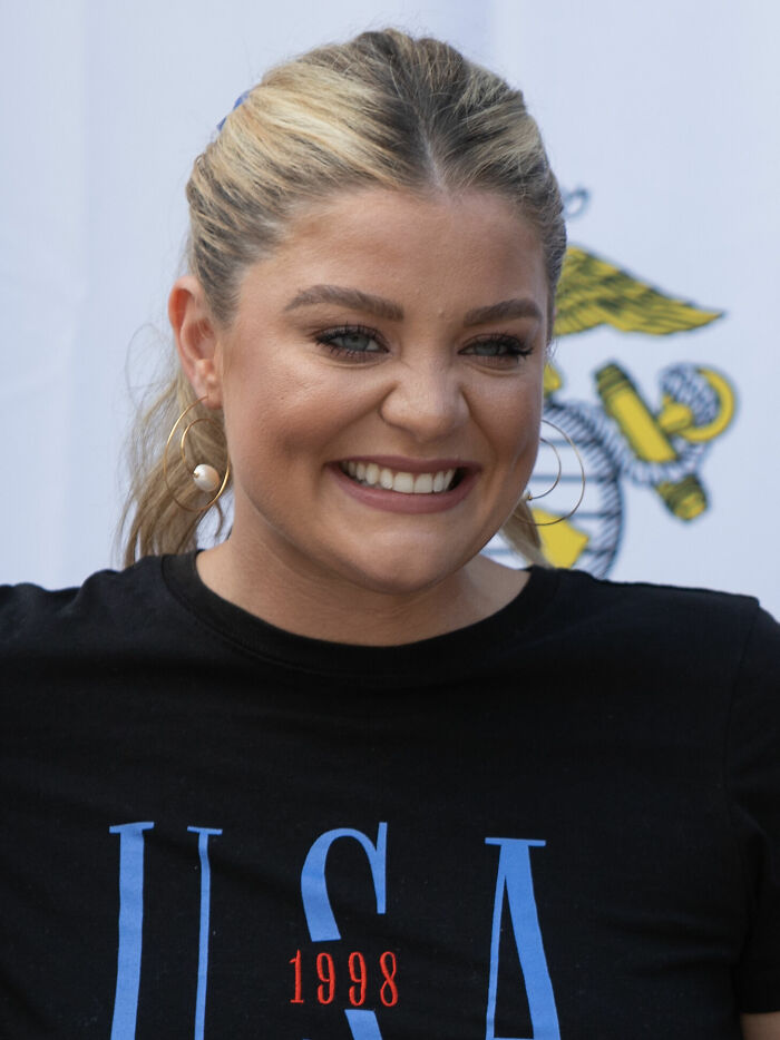Lauren Alaina smiling with hoop earrings and black USA shirt, attending an outdoor event with a patriotic backdrop.