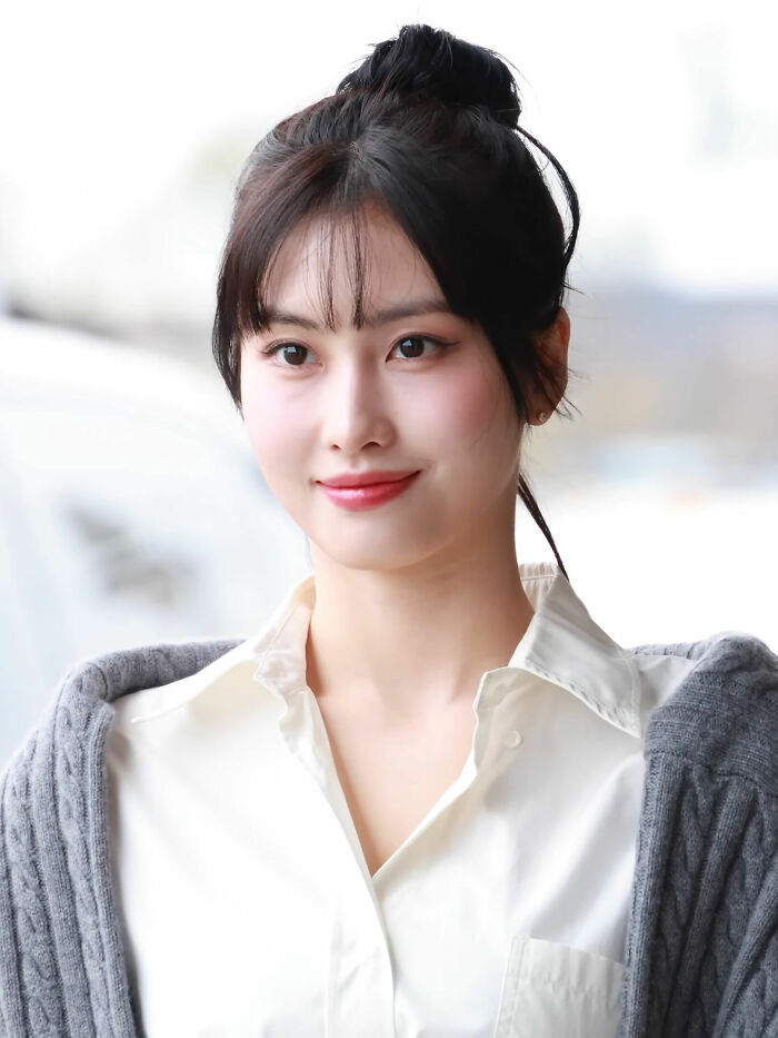 Momo Hirai smiling outdoors, wearing a white shirt and gray cardigan with her hair tied up in a bun.
