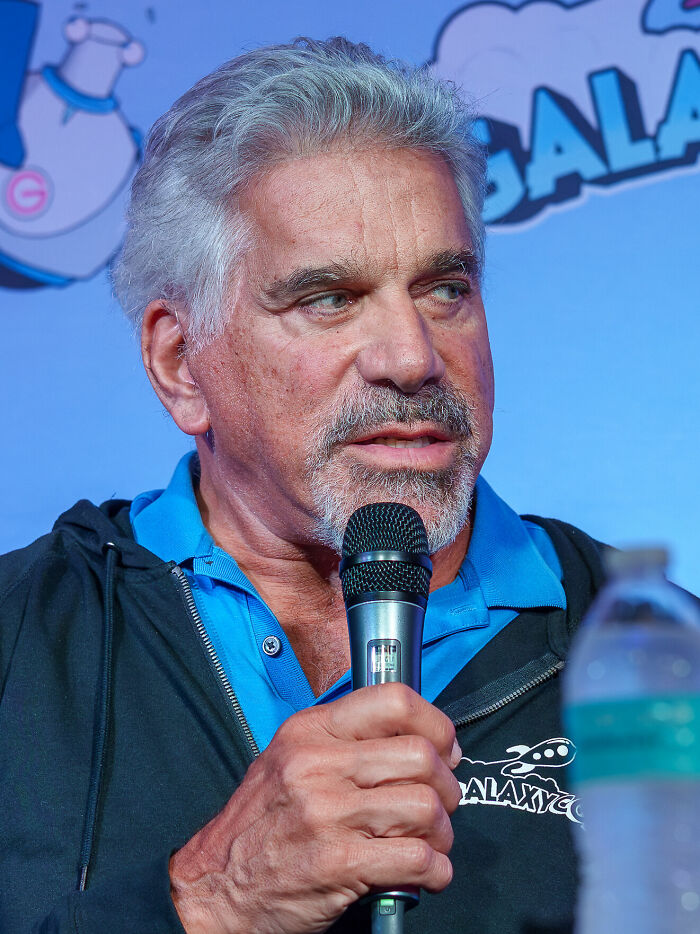 Lou Ferrigno speaking into a microphone at a public event, wearing a blue collared shirt and black jacket.