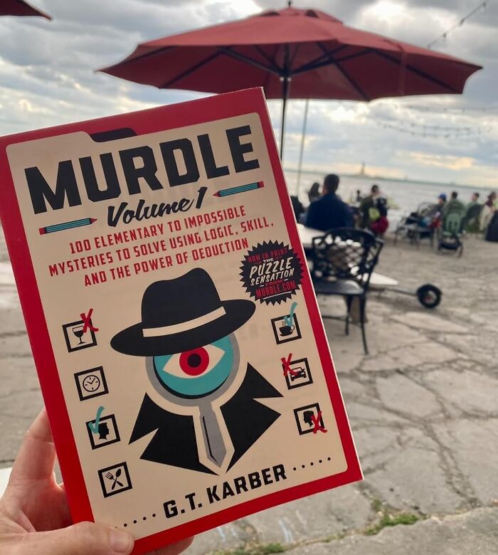 Hand holding Murdle puzzle book, a clever gift idea for people who are impossible to shop for, with outdoor seating in background.