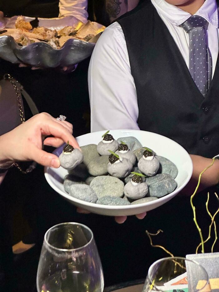 Hand holding oddly presented dish served on rocks at event, showcasing one of the dishes that are just stupid.