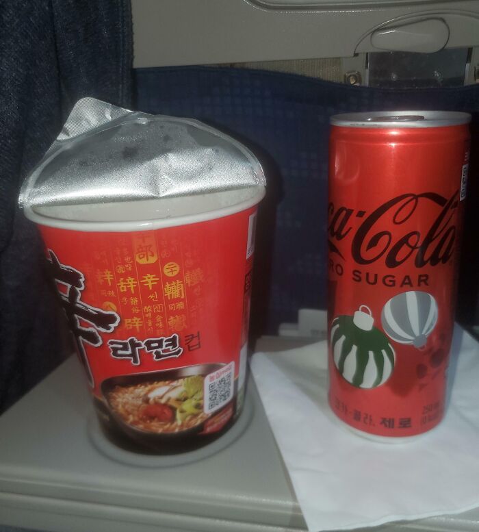 Unhinged pic of a cup of instant ramen with the lid sealed shut next to a Coca-Cola can on a tray table.