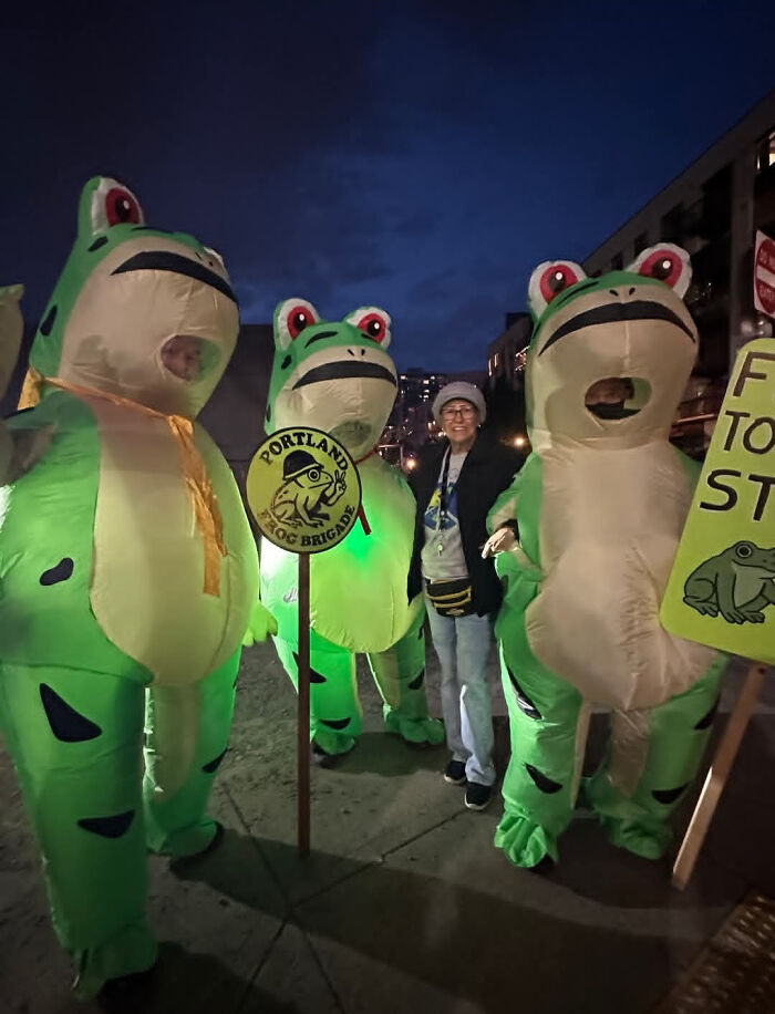 Three people in frog costumes standing outside at night with a woman holding a Portland Frog Brigade sign in a fail pics moment.