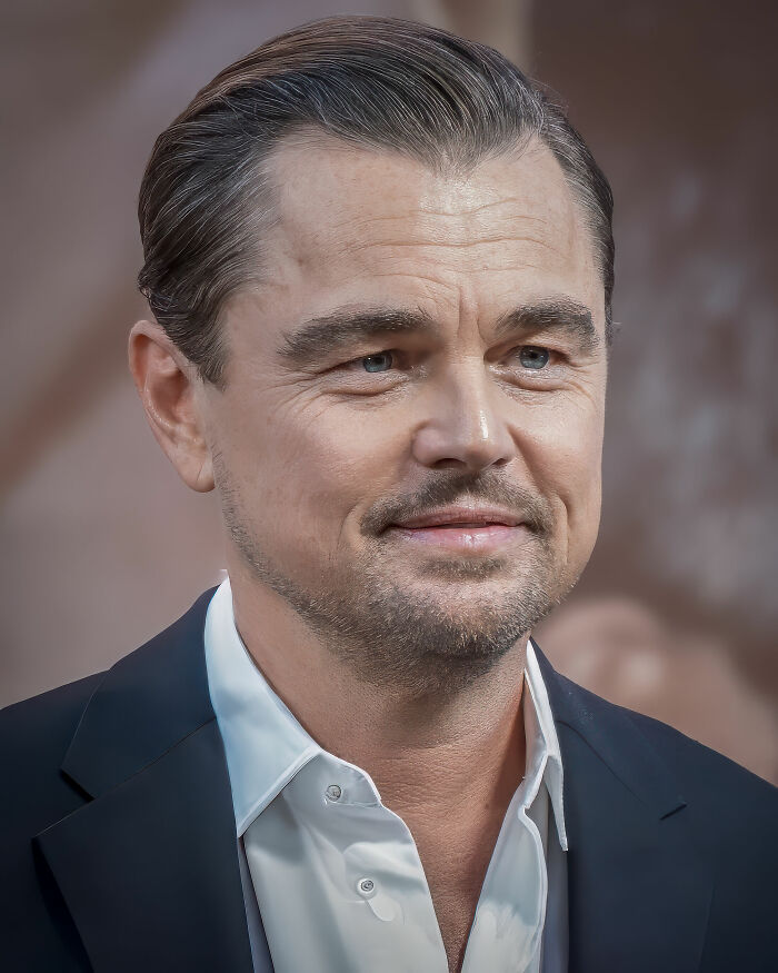 Leonardo DiCaprio close-up portrait with slicked-back hair, light beard, wearing a white shirt and dark blazer.