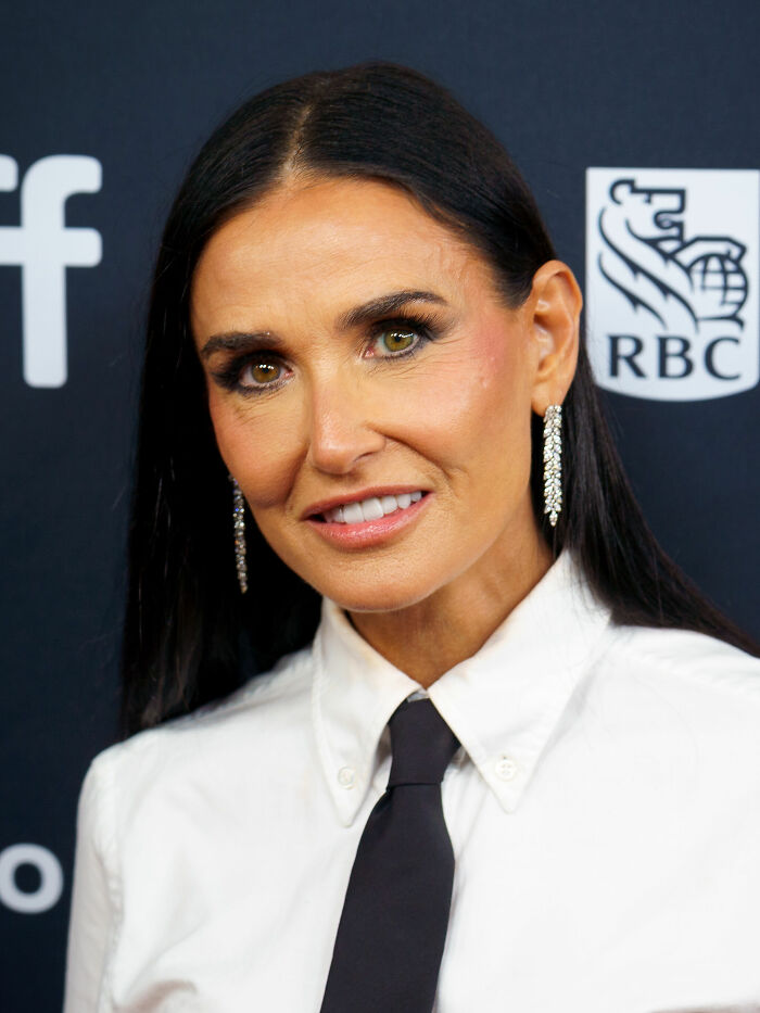 Demi Moore wearing white shirt and black tie with diamond earrings, smiling at a formal event background.