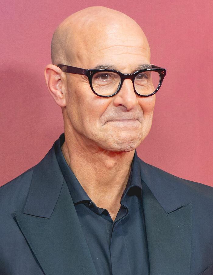 Stanley Tucci wearing black glasses and a dark suit, posing against a plain pink background in a close-up shot