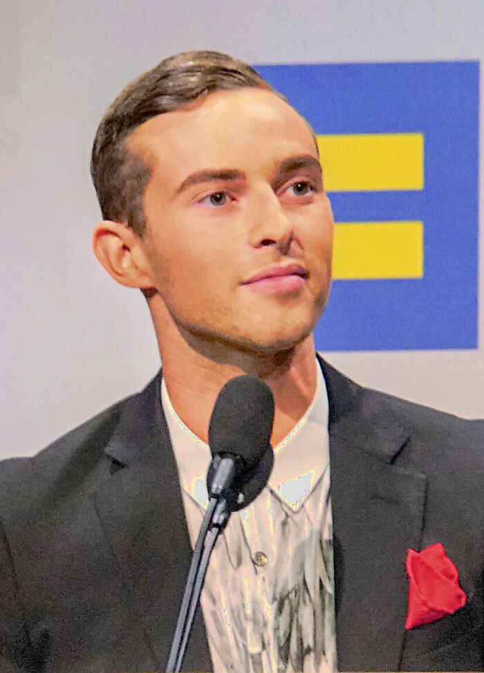 Adam Rippon speaking at an event, dressed formally with a microphone and a blue and yellow logo in the background