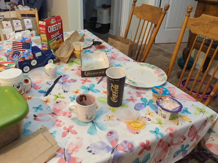 Cluttered family dining table with empty dishes, cereal box, drink cups, and scattered food items, showing family frustration.