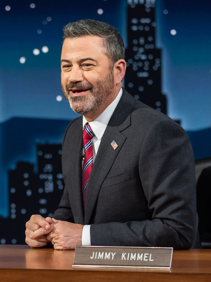 Jimmy Kimmel smiling in a suit and tie on a late night talk show set with a nameplate in front of him.