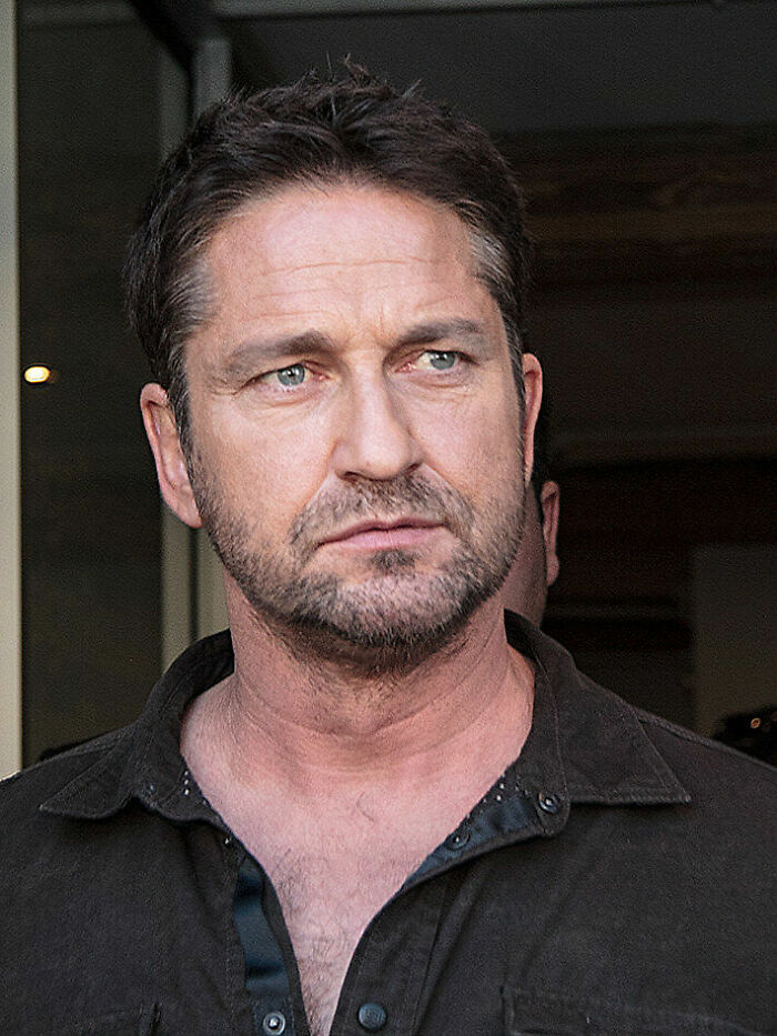 Gerard Butler with short hair and beard, wearing a dark shirt, looking serious in an indoor setting.