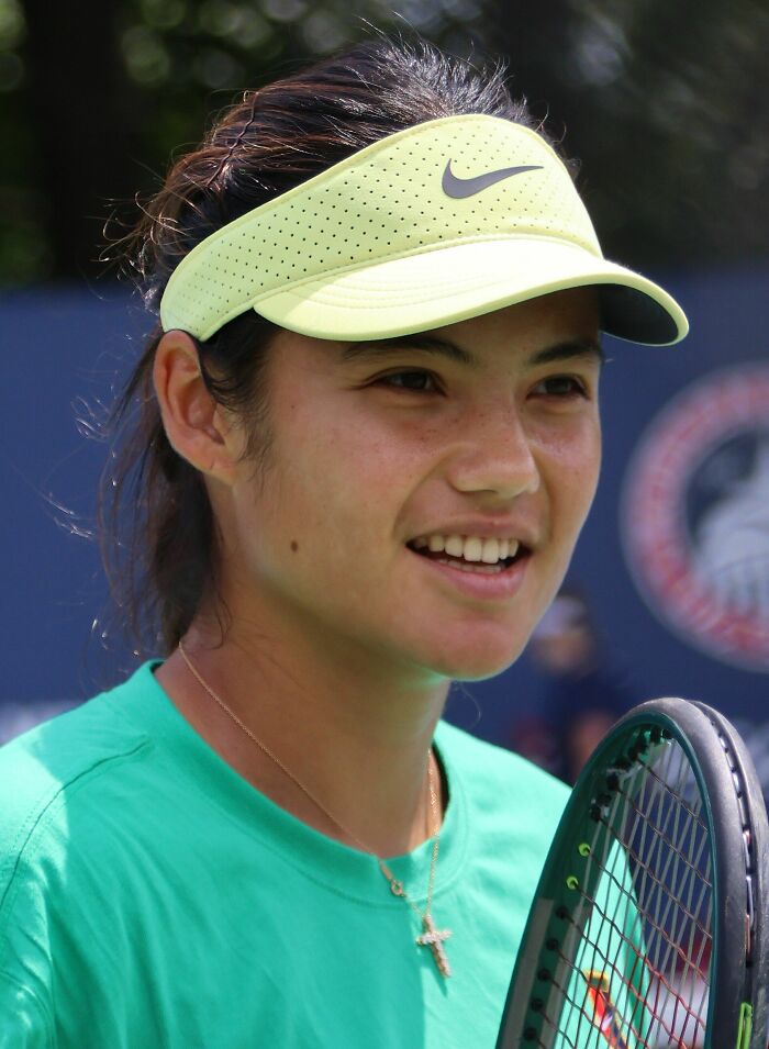 Emma Raducanu smiling during tennis practice, wearing a yellow visor and holding a tennis racket outdoors.