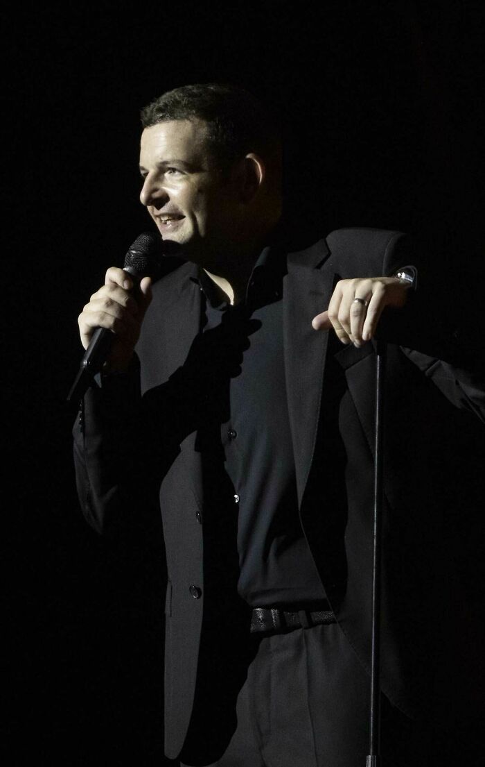 Kevin Bridges performing on stage, holding a microphone and leaning on a mic stand during a live comedy show.