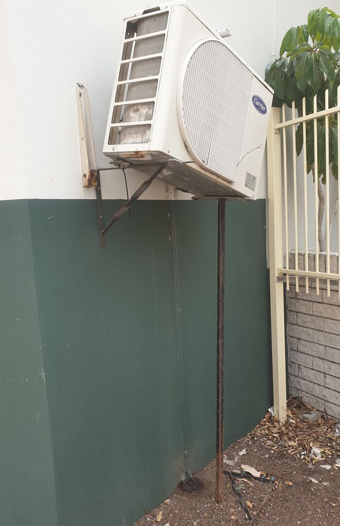 Old air conditioning unit propped up by a rusty metal pole showing a funny example of people not even trying.