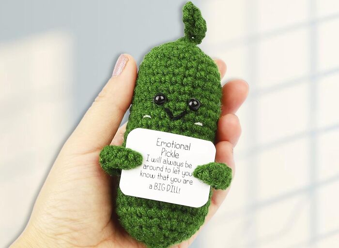 Hand holding a green crochet burr basket pickle plush with a cute face and a small note attached.