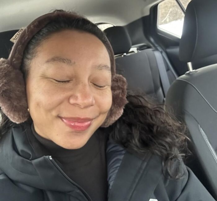 Woman wearing brown Burr Basket earmuffs and black jacket sitting inside a car, eyes closed and smiling peacefully.