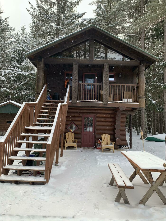 Cozy off-grid cabin in snowy forest with wooden stairs, balcony, outdoor chairs, and picnic table covered in snow.