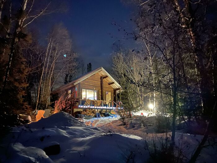 Cozy off-grid cabin lit warmly at night, surrounded by snow and bare trees in a peaceful winter forest setting.