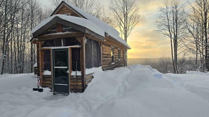 Cozy off-grid cabin covered in snow at sunset surrounded by bare trees in a peaceful winter landscape.