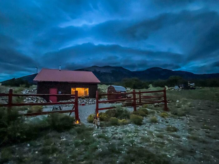 Cozy off-grid cabin with warm lights glowing at dusk, surrounded by a wooden fence and mountainous landscape.
