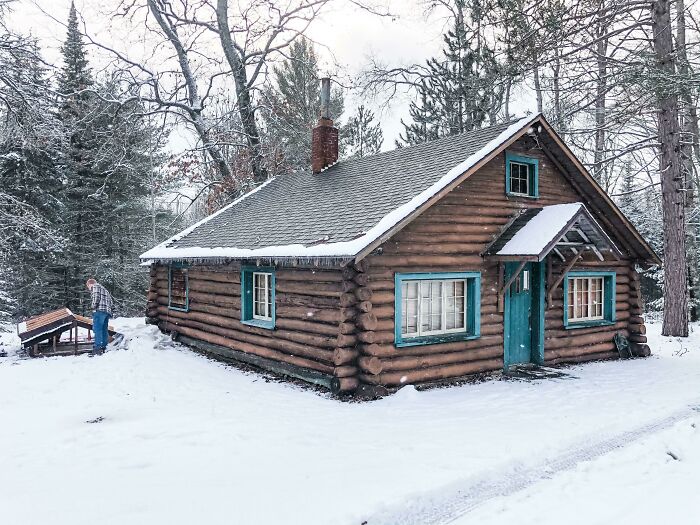 Cozy off-grid cabin in snowy forest setting with a person outside, surrounded by trees and winter landscape.