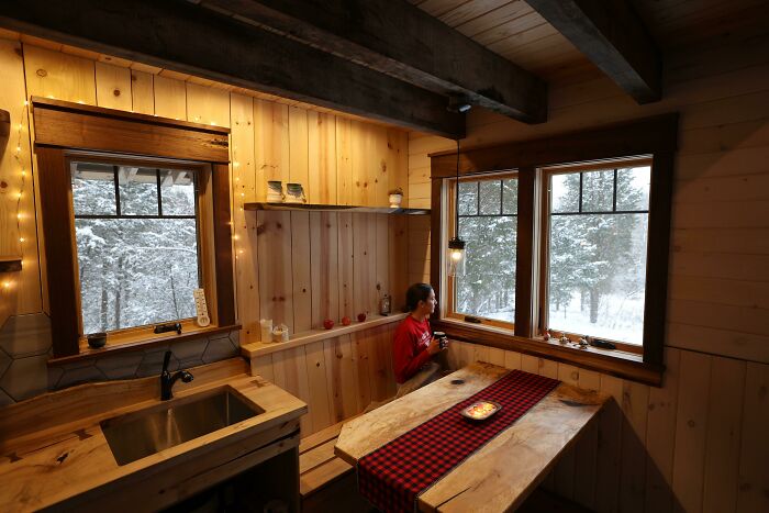 Cozy off-grid cabin interior with wooden walls, a woman looking out snowy windows, and warm ambient lighting.