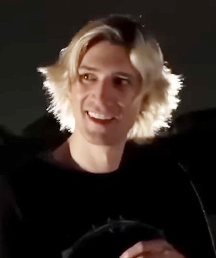 xQc smiling with blonde hair in a dark setting, highlighting moments from his bio and career highlights.