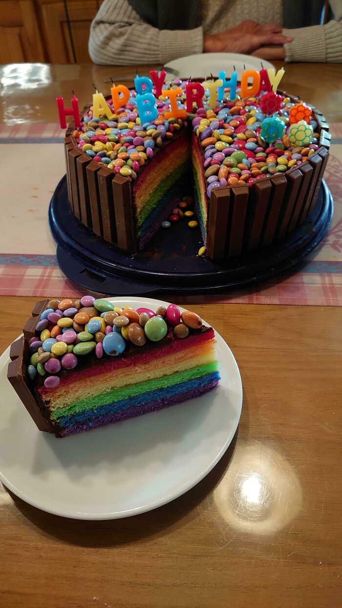 Rainbow layered birthday cake decorated with colorful candy and KitKat bars, celebrating a special sibling moment.