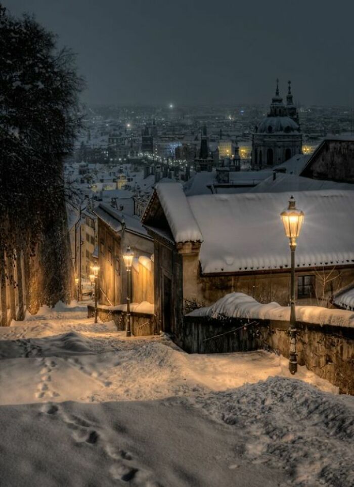 Snow-covered narrow street lit by warm lamps overlooking a cityscape at night, showing a different world perspective.