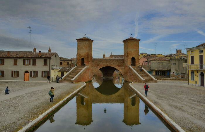Historic brick bridge with towers reflected in calm water, capturing our world from a unique perspective in an old town setting.