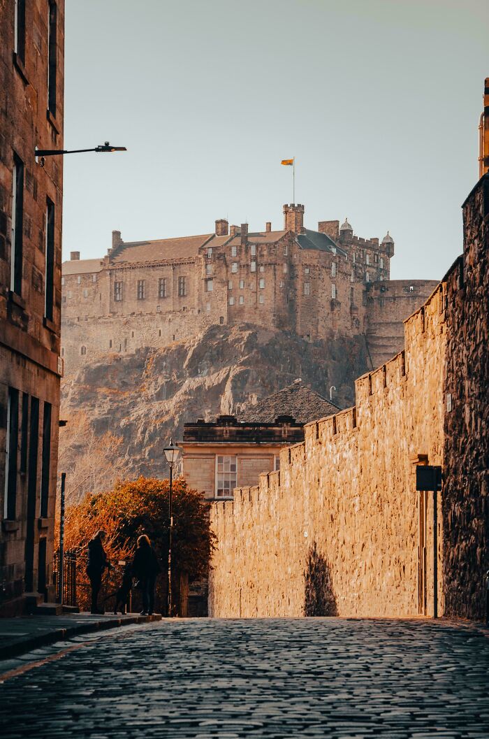 Historic castle atop rocky hill viewed from cobblestone street lined with stone walls, capturing unique world perspective.